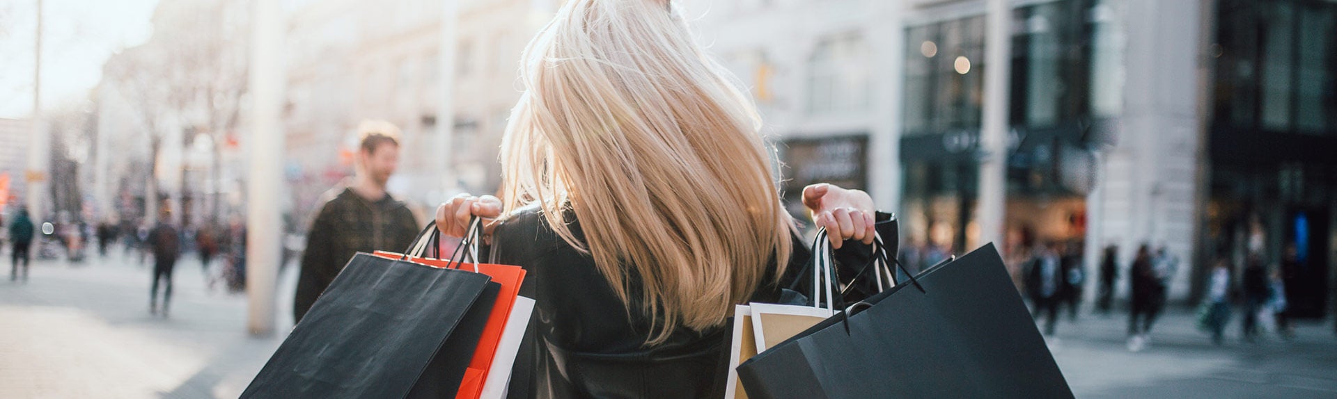 Image of back profile of woman walking down busy street carrying shopping bags in both hands.