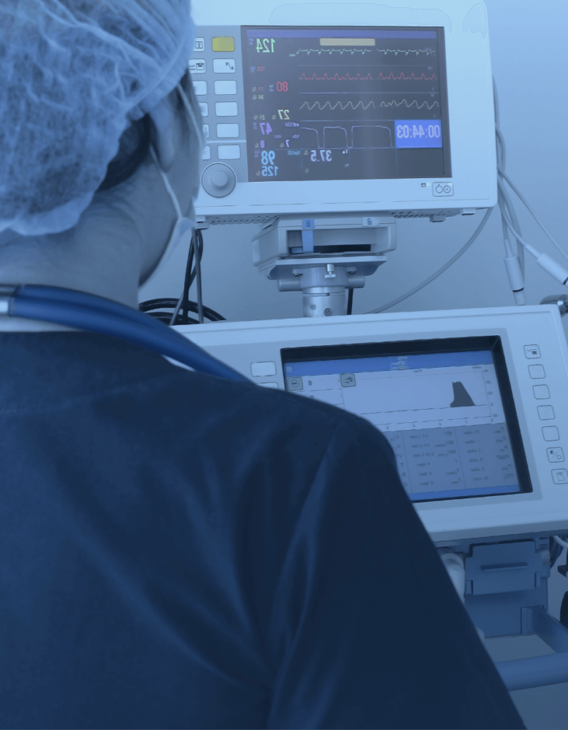 Lifestyle photo showing the back of a medical professional overlooking a patient monitoring machine.