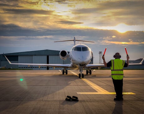 Photo showing the front view of a small commercial airplane on a runway at dawn with a marshaller standing in front guiding it where to park.