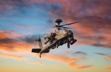 Photo of a Boeing AH-64 Apache helicopter flying through a partly cloudy sky during sunset.