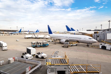 Photo showing a wide view of a jet bridge at the airport filled with parked commercial airplanes, baggage tugs, and ramp-ups.