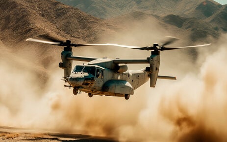 Photo of a Bell Boeing V-22 Osprey preparing to land atop a dirt field with dust kicking up and green hills in the background.