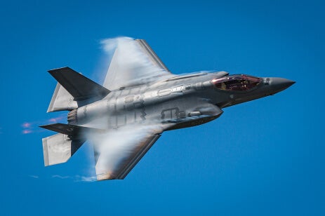 Photo of an F35 Falcon Fighter flying on its side against a clear blue sky.