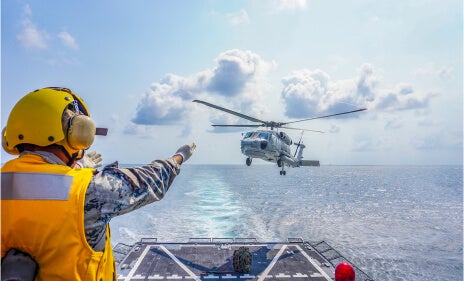 A photo of a military crew member on a ship at sea guiding a helicopter for landing on the ship.